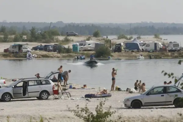 Ludzie odpoczywają nad jeziorem, niektórzy kąpią się, inni opalają na plaży. W tle widać kampery i samochody, a na wodzie pływa motorówka. To idealne miejsce na wakacje, jak na pogoria 4.