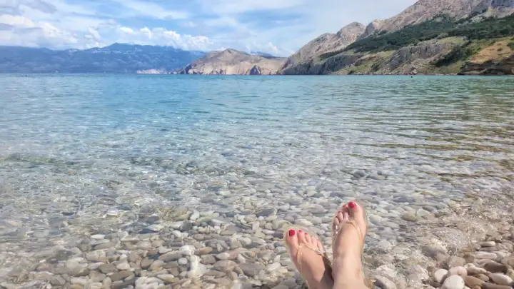 Stopy w klapkach na kamienistej plaży, krystalicznie czysta woda i górzyste wybrzeże. Raj na wyspie Krk.